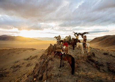Five eagle hunters carry their eagles on horseback over rugged cliffs just before the sunset.