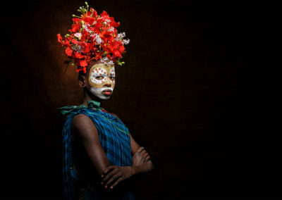 A young tribal girl from Ethiopia’s Omo Valley wears a red flower headdress and face paint, looking confidently into the camera.