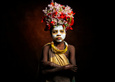 A young girl from Ethiopia’s Suri tribe stands with arms crossed, wearing a vibrant floral crown and face paint. Her traditional adornment includes natural elements like flowers and clay, reflecting the cultural artistry of the Omo Valley tribes.