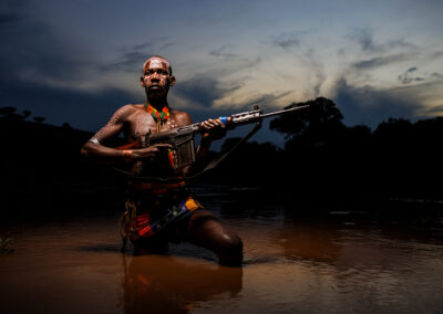 Portrait of a Karo tribe man in the Omo Valley, Ethiopia, holding a Kalashnikov rifle in a river, with ceremonial body paint.