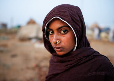 Young girl in a maroon shawl with a gold nose ring looks intensely into the camera at the Pushkar Camel Fair grounds in Rajasthan, India, early morning.