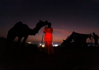Silhouette of a camel leaning toward its owner under a purple night sky at Pushkar Mela, with soft red light illuminating the figures and distant fairground lights in the background.