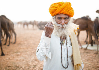 Elderly Rajasthani camel trader with an orange turban and white beard standing among camels at the Pushkar Mela.