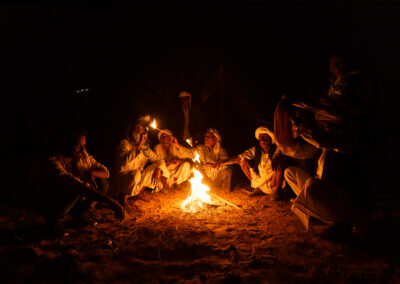 Group of men in traditional Rajasthani attire sitting around a small bonfire at night, their faces lit warmly by the flames.