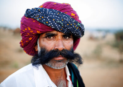 Close-up portrait of a Rajasthani camel trader wearing a red-and-black turban with a large moustache at the Pushkar fair.