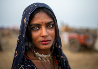 Portrait of a young Rajasthani woman with striking light brown eyes, a navy blue printed headscarf, nose stud, and traditional necklace, looking directly at the camera at Pushkar Mela.