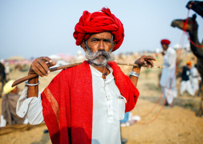 Portrait of a Rajasthani man with a red turban and shawl, holding a wooden stick across his shoulders at the Pushkar camel fair.