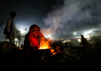 Rajasthani woman blowing air into a small bonfire at night to cook food during Pushkar Mela, her face lit by the warm glow of the fire.