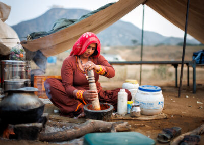 A woman in traditional Rajasthani attire sits cross-legged under a tent at Pushkar Mela, grinding spices in a stone mortar with cooking utensils and food supplies arranged around her.